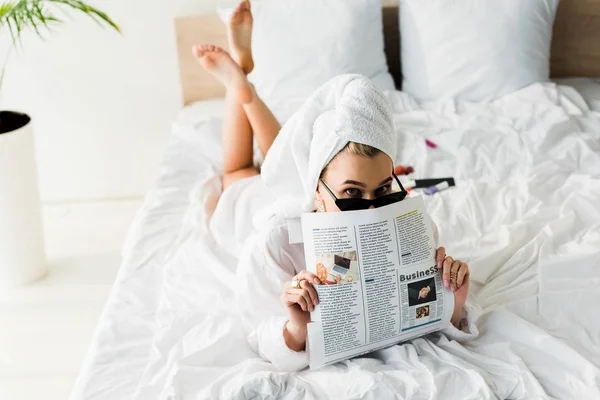 Elegante donna a piedi nudi in camicia, occhiali da sole, gioielli e con asciugamano sulla testa leggendo giornale business a letto — Foto stock