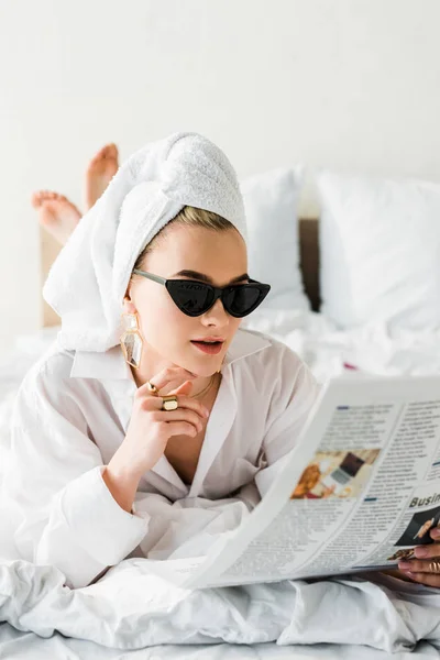 Foco seletivo da mulher descalça elegante na camisa, óculos de sol, jóias e com toalha na cabeça ler jornal na cama — Fotografia de Stock