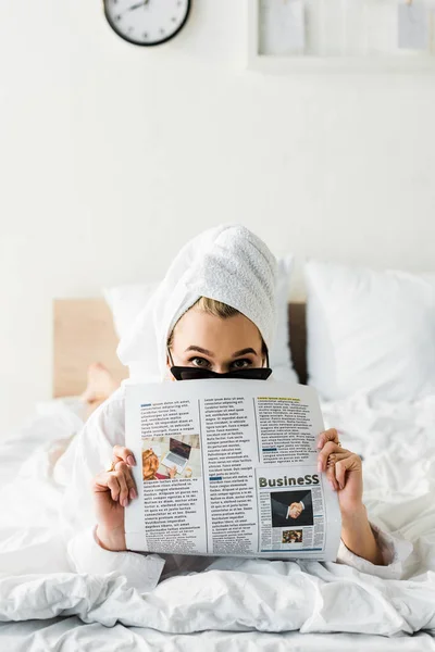 Mulher em óculos de sol, jóias e com toalha na cabeça lendo jornal de negócios na cama — Fotografia de Stock