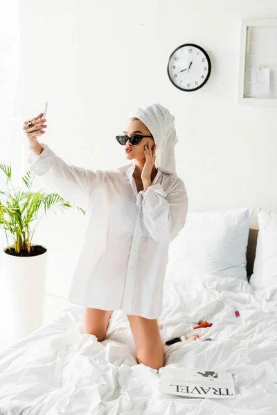 Mujer con estilo en camisa, gafas de sol, joyas y con toalla en la cabeza tomando selfie en la cama - foto de stock