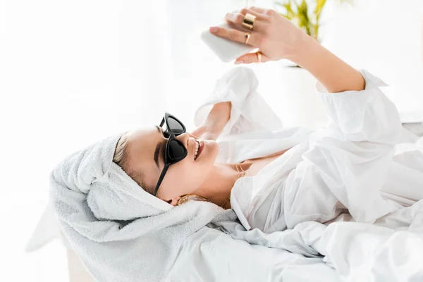 Joven mujer sonriente en camisa, gafas de sol, joyas y con toalla en la cabeza tomando selfie en el teléfono inteligente en la cama - foto de stock