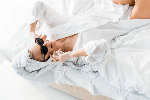 Молода усміхнена жінка в сорочці, сонцезахисних окулярах, прикрасах і з рушником на голові розмовляє на смартфоні в ліжку — стокове фото