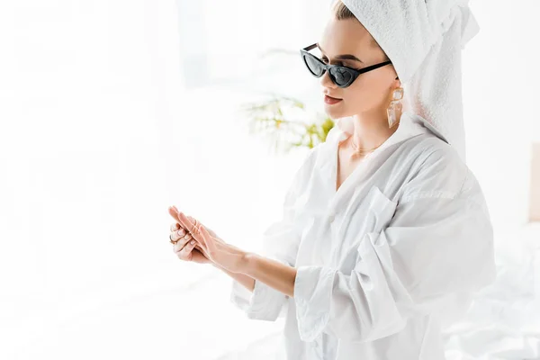 Young stylish woman in shirt, sunglasses, jewelry and with towel on head looking at hand — Stock Photo