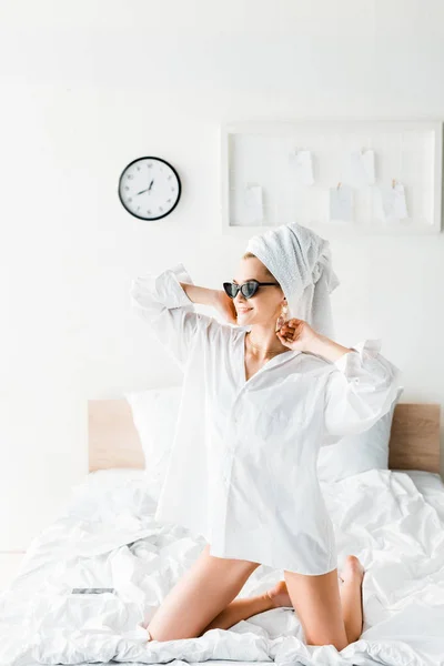 Mujer joven y elegante feliz en camisa, gafas de sol, joyas y con toalla en la cabeza sentado en la cama - foto de stock