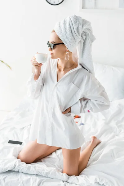 Jeune femme élégante en chemise, lunettes de soleil, bijoux et avec serviette sur la tête assise sur le lit tout en buvant du café — Photo de stock