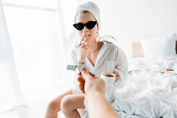 Young stylish woman in shirt, jewelry and sunglasses with towel on head and cup of coffee lighting up cigarette while sitting on bed — Stock Photo