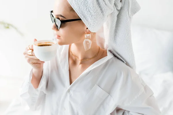 Jeune femme élégante en chemise, lunettes de soleil, bijoux et avec serviette sur la tête boire du café — Photo de stock