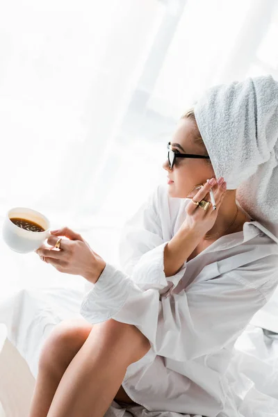 Mujer joven y elegante en joyas y gafas de sol con toalla en la cabeza y taza de café fumar cigarrillo - foto de stock