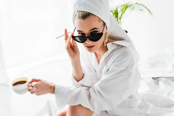 Junge stilvolle Frau in Schmuck und Sonnenbrille mit Handtuch auf dem Kopf und einer Tasse Kaffee, Zigarette rauchend und in die Kamera blickend — Stockfoto
