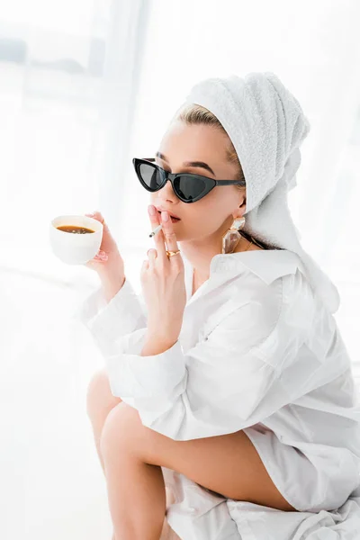 Jeune femme élégante en bijoux et lunettes de soleil avec serviette sur la tête et tasse de café cigarette fumeur le matin — Photo de stock