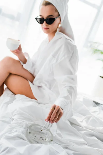 Selektiver Fokus der jungen stilvollen Frau in Schmuck und Sonnenbrille mit Handtuch auf dem Kopf und Tasse Kaffee rauchende Zigarette mit Aschenbecher auf dem Bett — Stockfoto