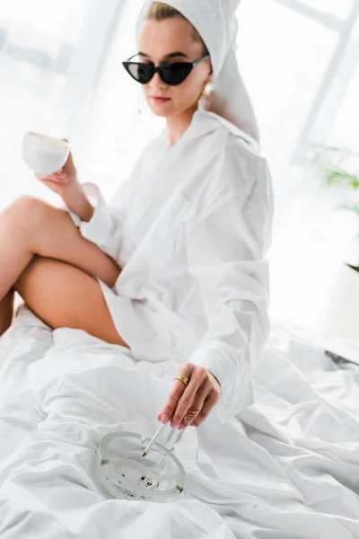 Selective focus of young stylish woman in jewelry and sunglasses with towel on head and cup of coffee smoking cigarette with ashtray on bed — Stock Photo