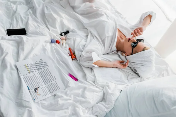 Vue aérienne de la jeune femme élégante en bijoux et lunettes de soleil avec serviette sur la tête couchée dans le lit près de vernis à ongles, lime à ongles, smartphone et journal — Photo de stock