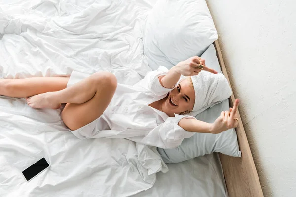 Вид зверху босоніж щаслива жінка в прикрасах з рушником на голові лежить у білому ліжку біля смартфона і показує середні пальці — стокове фото