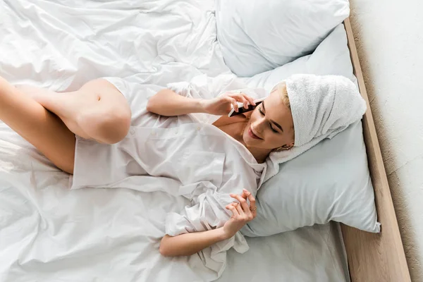 Top view of smiling woman in jewelry with towel on head lying in white bed and talking on smartphone — Stock Photo