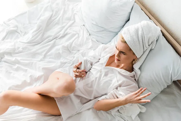 Vista superior de la mujer insatisfecha en joyas con toalla en la cabeza acostado en la cama blanca y el uso de teléfono inteligente - foto de stock