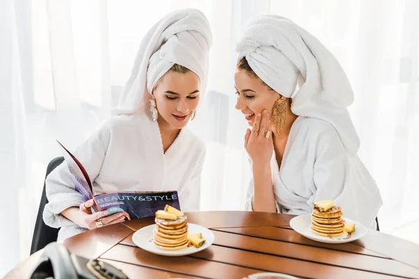 Élégantes femmes souriantes en peignoirs et bijoux avec des serviettes sur les têtes lecture magazine ensemble près de la table avec des crêpes — Photo de stock