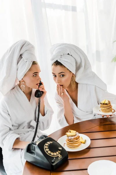 Mujeres impactadas con estilo en albornoces y joyas con toallas en las cabezas hablando por teléfono retro mientras desayunaba - foto de stock
