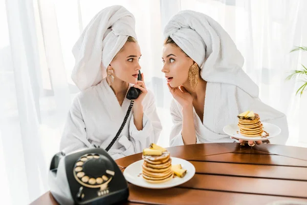 Mujeres impactadas con estilo en albornoces y joyas con toallas en las cabezas hablando por teléfono retro mientras desayunan y se miran entre sí - foto de stock