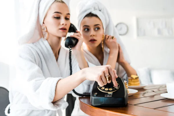 Enfoque selectivo de las mujeres con estilo en batas de baño y joyas con toallas en las cabezas utilizando el teléfono retro mientras está sentado en la mesa - foto de stock