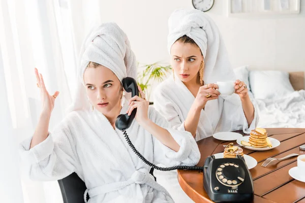 Mulheres elegantes em roupões de banho e jóias com toalhas na cabeça usando telefone retro enquanto tomam café da manhã juntos — Fotografia de Stock