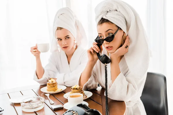 Elegante donna in accappatoio, occhiali da sole e gioielli con asciugamano in testa utilizzando telefono retrò mentre fa colazione con un amico — Foto stock