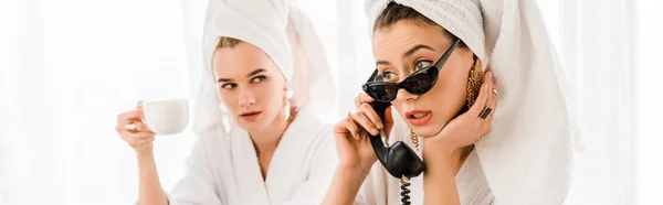 Панорамний знімок стильної жінки в халаті, сонцезахисних окулярах та прикрасах з рушником на голові, використовуючи ретро телефон біля жінки з чашкою — стокове фото