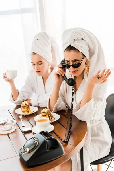 Mulher insatisfeita elegante em roupão de banho, óculos de sol e jóias com toalha na cabeça falando no telefone retro enquanto toma café da manhã com o amigo — Fotografia de Stock