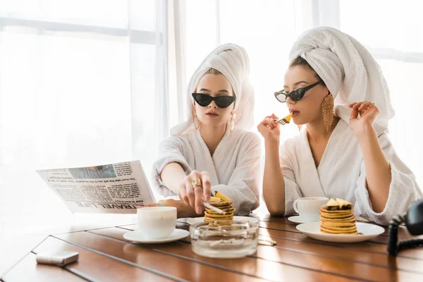 Stylish women in bathrobes, sunglasses and jewelry with towels on heads smoking cigarette and reading newspaper while eating pancakes — Stock Photo