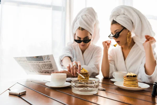 Selective focus of stylish women in bathrobes, sunglasses and jewelry with towels on heads smoking cigarette and reading newspaper while eating pancakes — Stock Photo