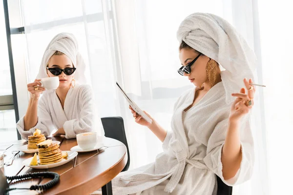 Stylish women in bathrobes, sunglasses and jewelry with towels on heads smoking cigarette and reading newspaper at morning — Stock Photo