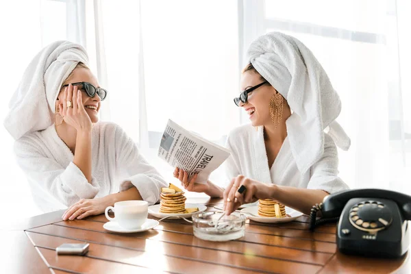 Mulheres felizes elegantes em roupões de banho, óculos de sol e jóias com toalhas na cabeça fumando cigarro, rindo e lendo jornal pela manhã — Fotografia de Stock