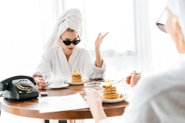 Mujer con estilo insatisfecho en albornoz, gafas de sol y joyas con toalla en la cabeza hablando con un amigo durante el desayuno - foto de stock