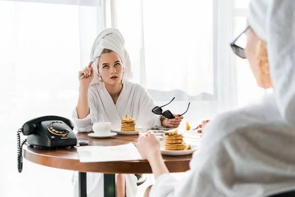 Mujer con estilo insatisfecho en albornoz y joyas con toalla en la cabeza y gafas de sol rodando los ojos durante el desayuno con un amigo - foto de stock