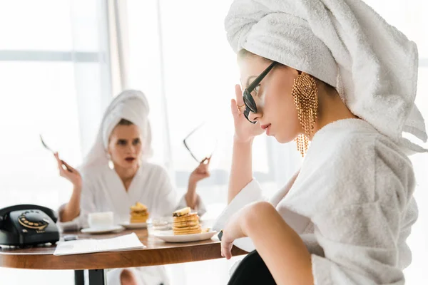 Enfoque selectivo de las mujeres con estilo en batas de baño y joyas con toallas en las cabezas peleando durante el desayuno - foto de stock