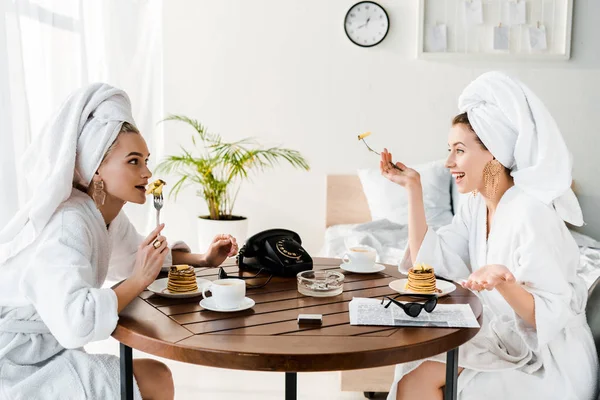 Élégantes femmes heureuses en peignoirs et bijoux avec des serviettes sur les têtes parler pendant le petit déjeuner — Photo de stock
