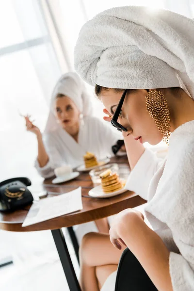 Selective focus of upset stylish woman in bathrobe, sunglasses and jewelry with towel on head sitting at table — Stock Photo