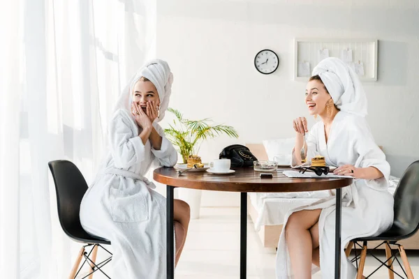 Mujeres elegantes felices y conmocionadas en batas de baño y joyas con toallas en las cabezas desayunando - foto de stock