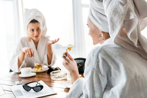 Вибірковий фокус усміхненої стильної жінки в халаті та прикрасах з рушником на голові, сидячи за столом і розмовляючи з другом — стокове фото