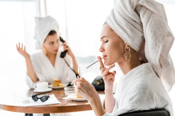 Enfoque selectivo de la mujer con estilo en el albornoz y la joyería con toalla en la cabeza iluminando el cigarrillo mientras que el amigo habla por teléfono - foto de stock