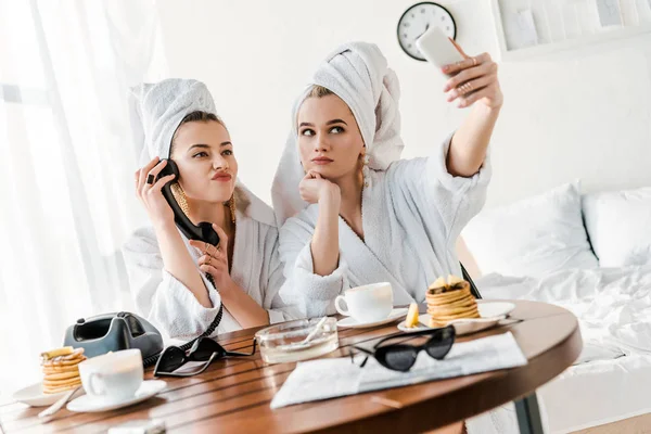 Foco seletivo de mulheres elegantes em roupões de banho e jóias com toalhas na cabeça falando no telefone retro e sorrindo ao tomar selfie — Fotografia de Stock