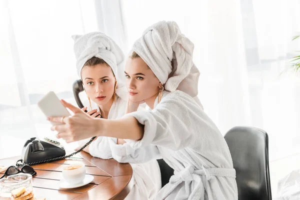 Selective focus of stylish women in bathrobes and jewelry with towels on heads talking on retro phone and taking selfie — Stock Photo