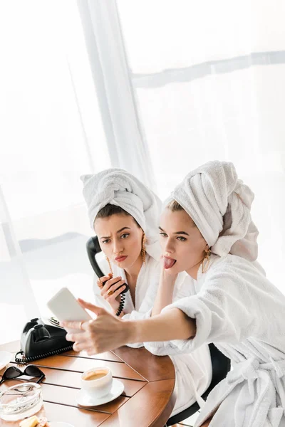 Selective focus of stylish women in bathrobes and jewelry with towels on heads talking on retro phone and grimacing while taking selfie on smartphone — Stock Photo