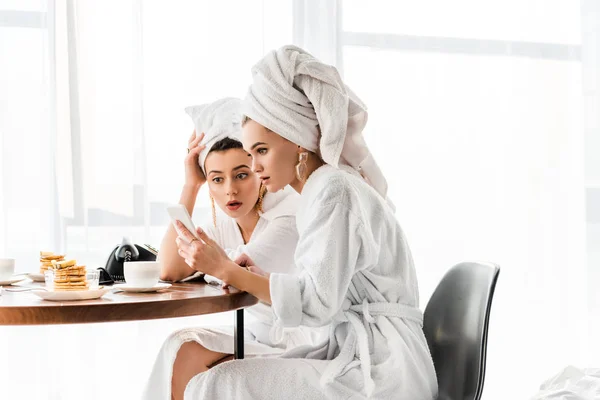 Mujeres impactadas con estilo en batas de baño y joyas con toallas en las cabezas utilizando el teléfono inteligente durante el desayuno - foto de stock