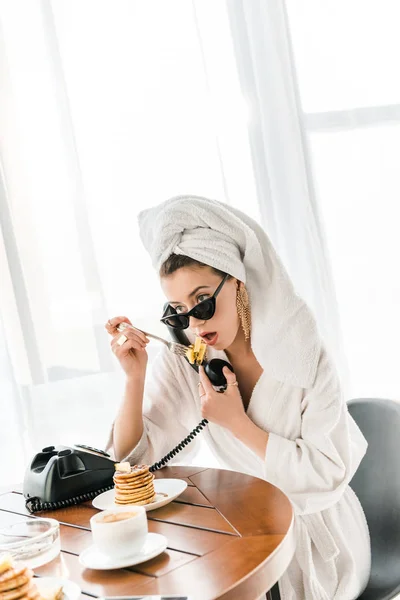 Mujer elegante en albornoz, gafas de sol y joyas con toalla en la cabeza hablando por teléfono vintage mientras come panqueques - foto de stock