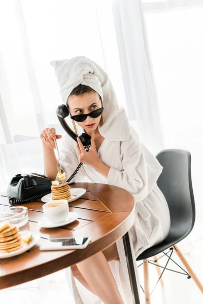 Mulher elegante em roupão de banho, óculos de sol e jóias com toalha na cabeça falando no telefone retro enquanto come panquecas — Fotografia de Stock