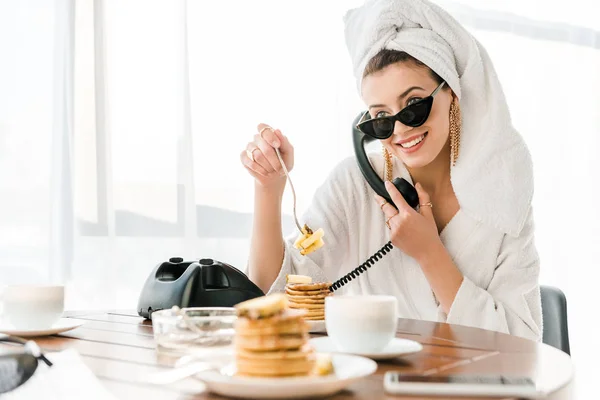 Elegante donna sorridente in accappatoio, occhiali da sole e gioielli con asciugamano in testa che parla al telefono retrò mentre mangia frittelle — Foto stock