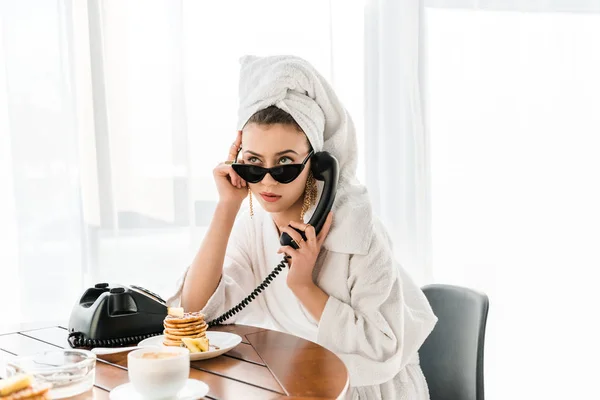 Стильна жінка в халаті, сонцезахисних окулярах та ювелірних виробах з рушником на голові розмовляє на ретро телефоні під час сніданку — стокове фото
