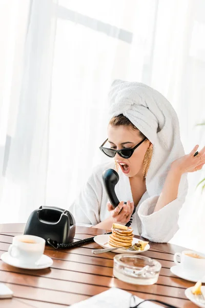 Mulher elegante irritado em roupão de banho, óculos de sol e jóias com toalha na cabeça gritando enquanto fala no telefone retro — Fotografia de Stock