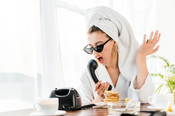 Mujer con estilo irritado en albornoz, gafas de sol y joyas con toalla en la cabeza gritando y haciendo gestos mientras habla por teléfono retro - foto de stock